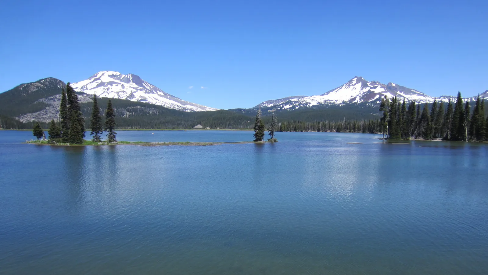 Calm blue water on Sparks Lake with small tree-covered islands and South Sister and Broken Top behind