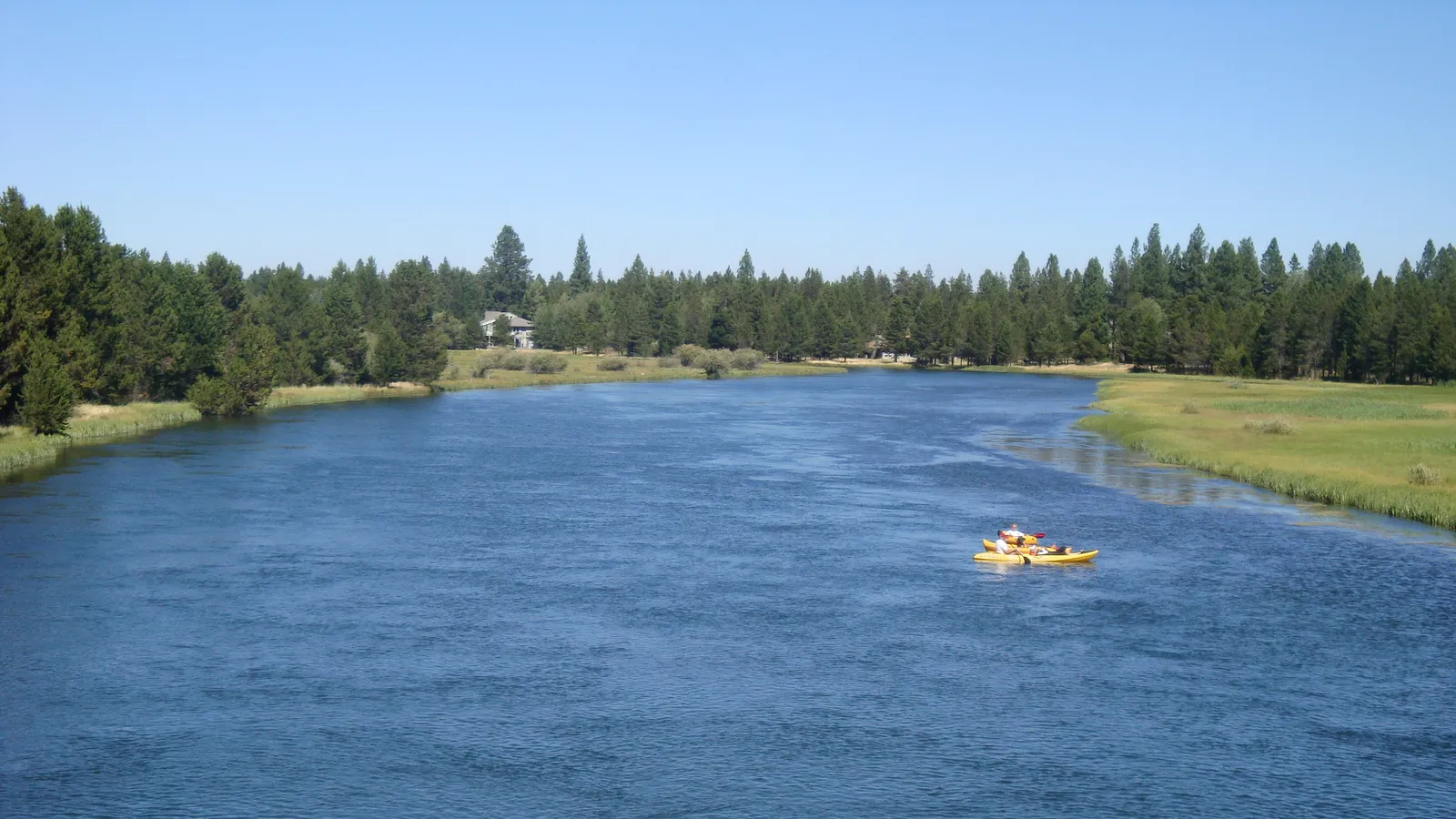 A kayaker on the wide, calm Deschutes River at Sunriver, lined with pines and meadow