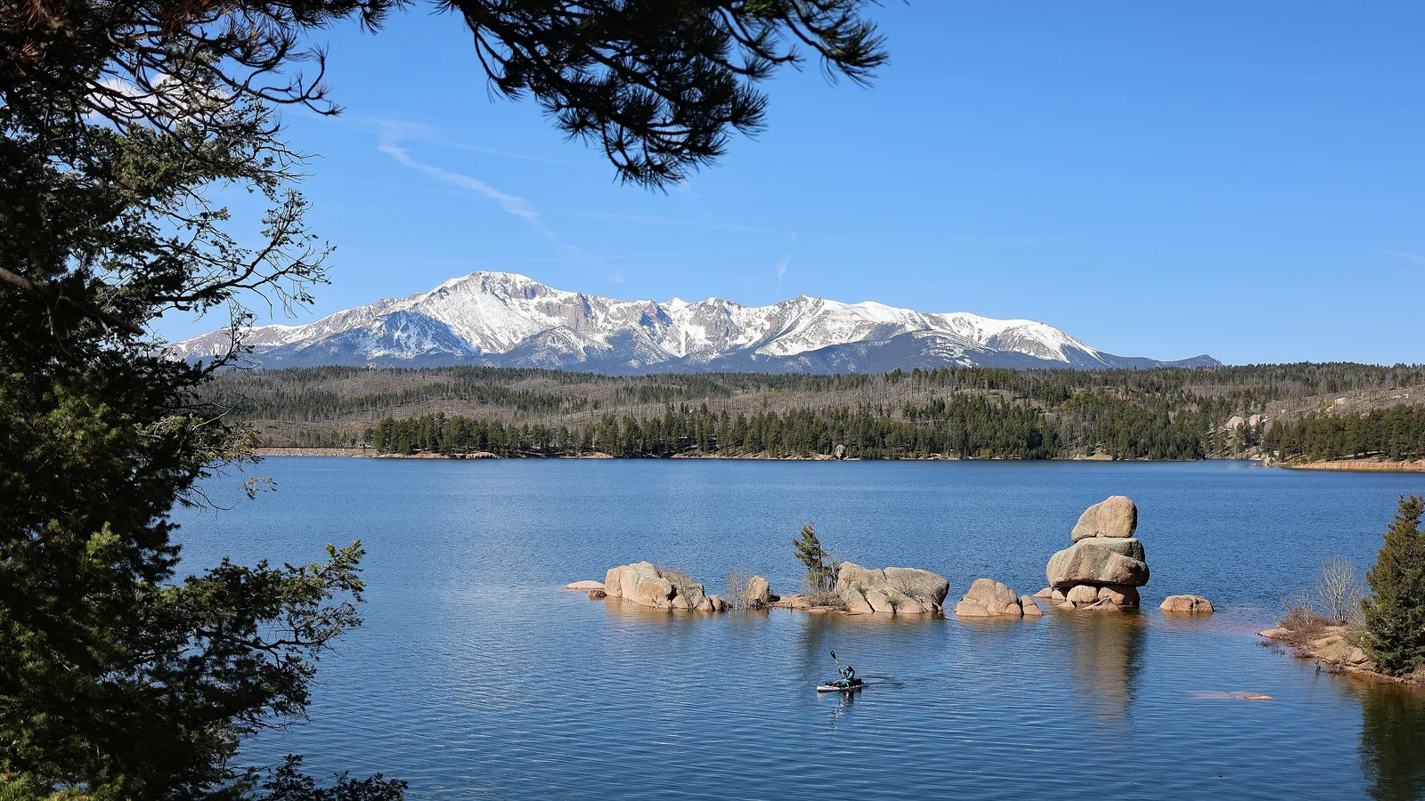 A kayaker among granite boulders on Rampart Reservoir, with snowy Pikes Peak in the distance