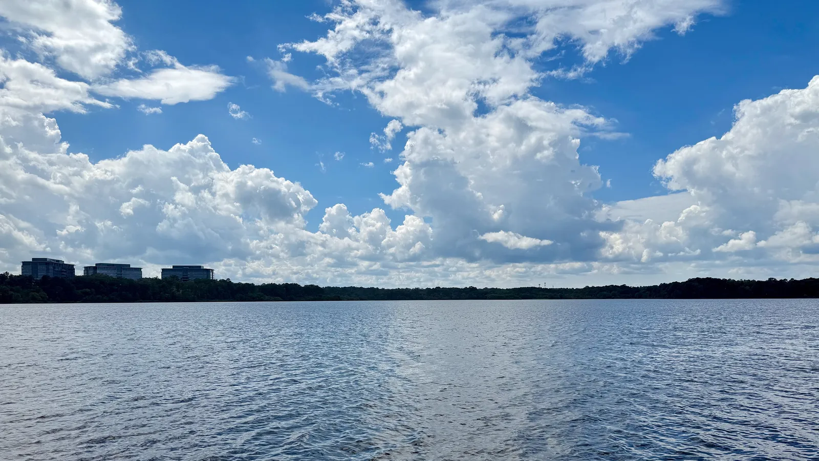 Open water on Lake Crabtree under summer clouds, with office buildings on the far shore
